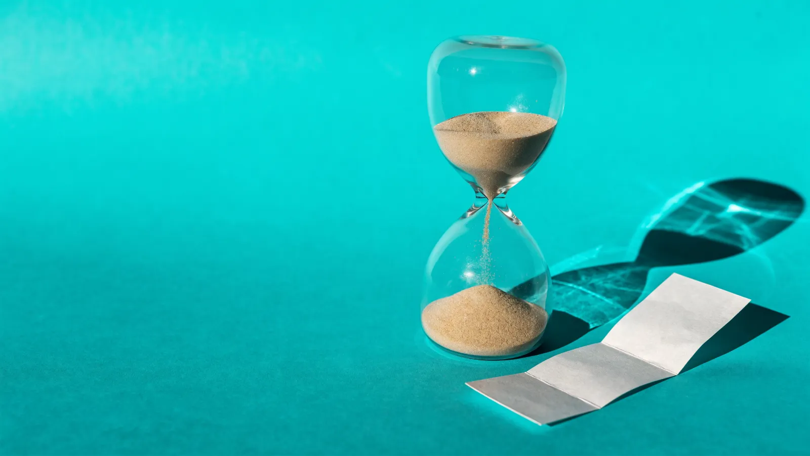 Un reloj de arena con casi toda la arena abajo junto a una etiqueta de papel en blanco, sobre fondo turquesa