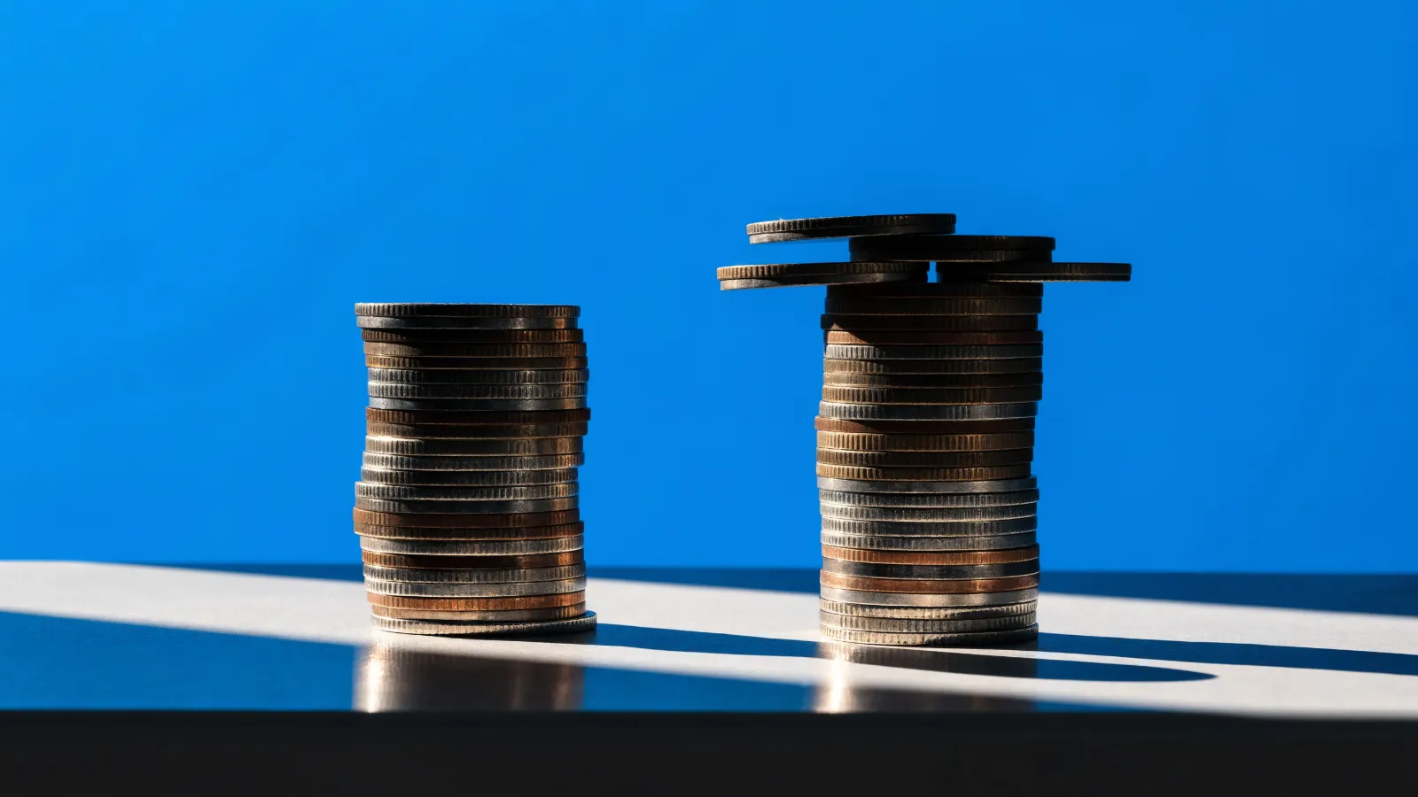 Dos pilas de monedas iguales, una con tres monedas extra encima, sobre fondo azul
