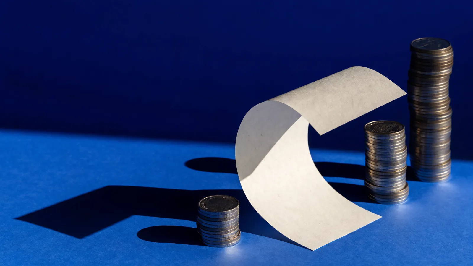 Un ticket de papel en blanco junto a tres pilas de monedas de distinta altura, sobre fondo azul