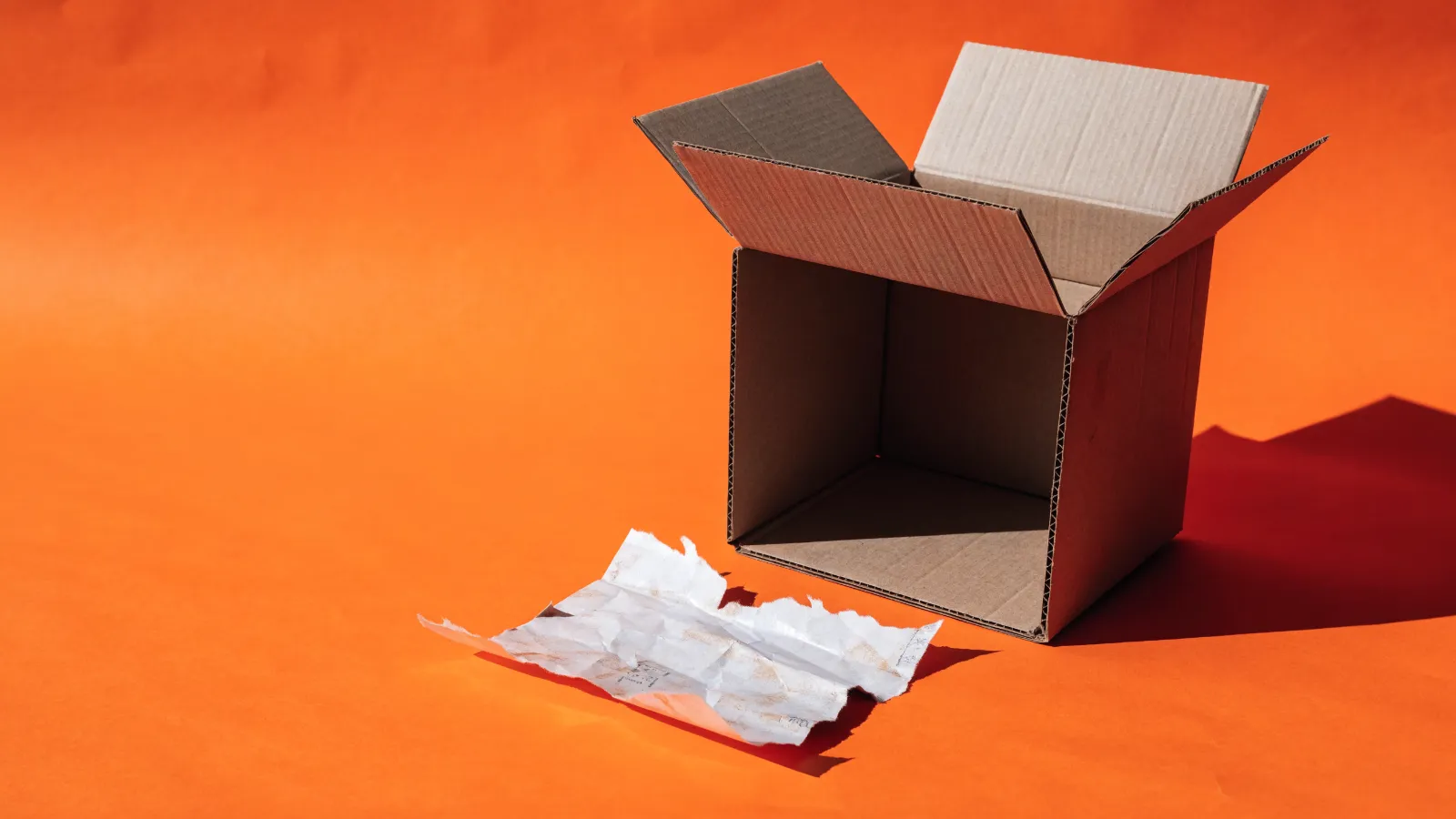 Una caja de envío abierta y vacía junto a una guía de envío rota, sobre fondo naranja
