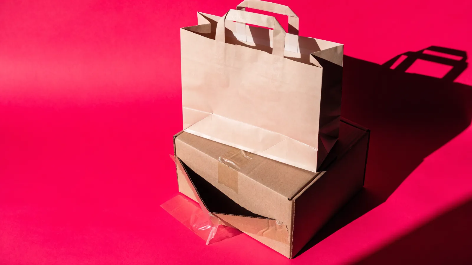 Una bolsa de compras nueva sobre una caja de envío con una esquina aplastada, sobre fondo rosa