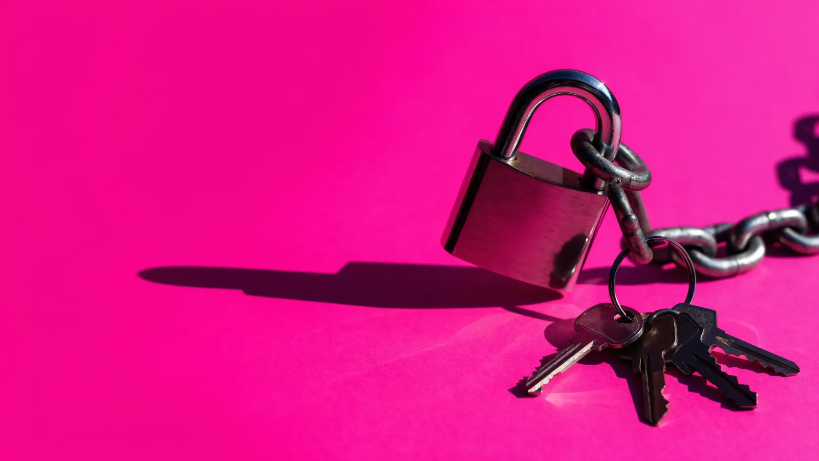 Un candado nuevo cerrado sobre una cadena cuya única llave cuelga de un manojo ajeno, sobre fondo rosa
