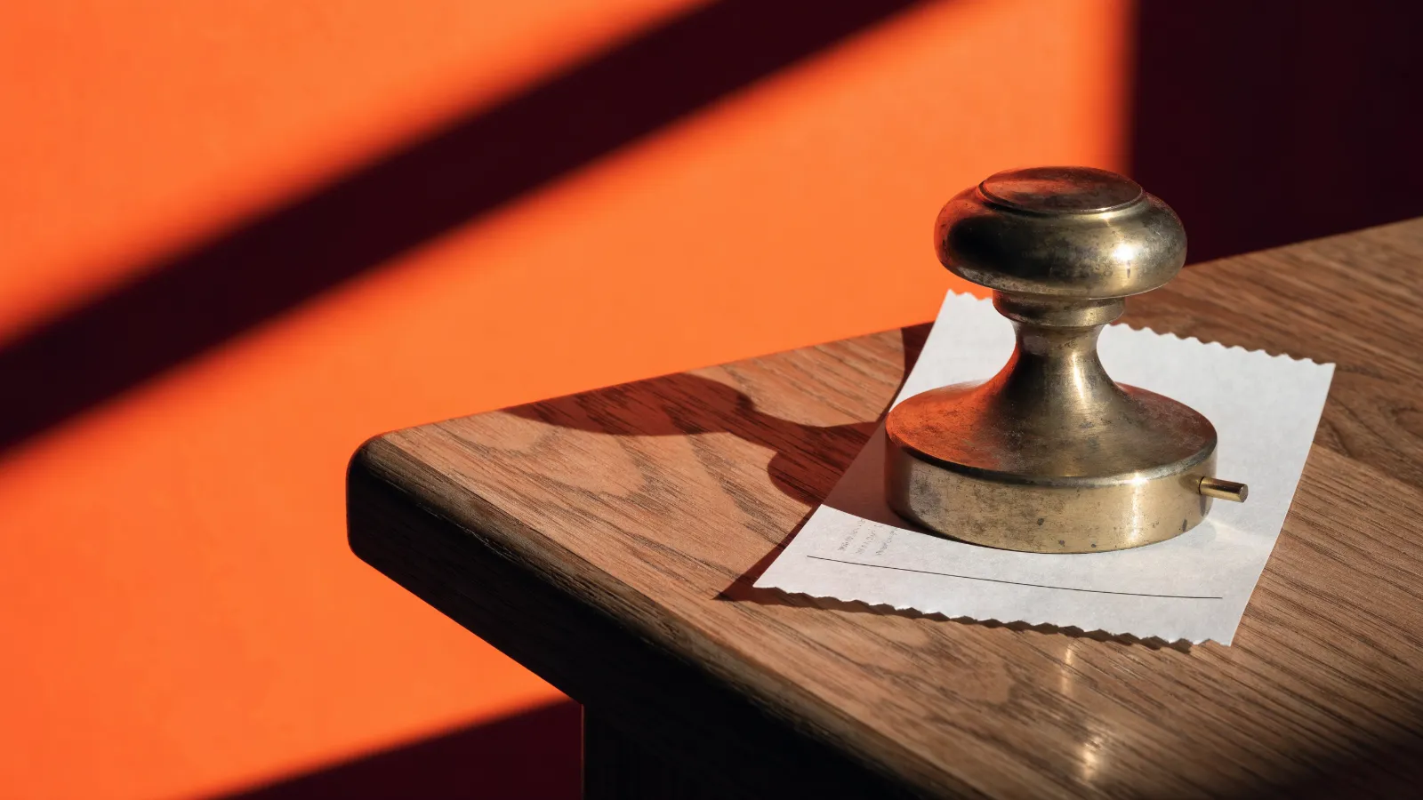 Un ticket de venta prensado bajo un sello de banco de latón sobre un escritorio de madera, sobre fondo naranja