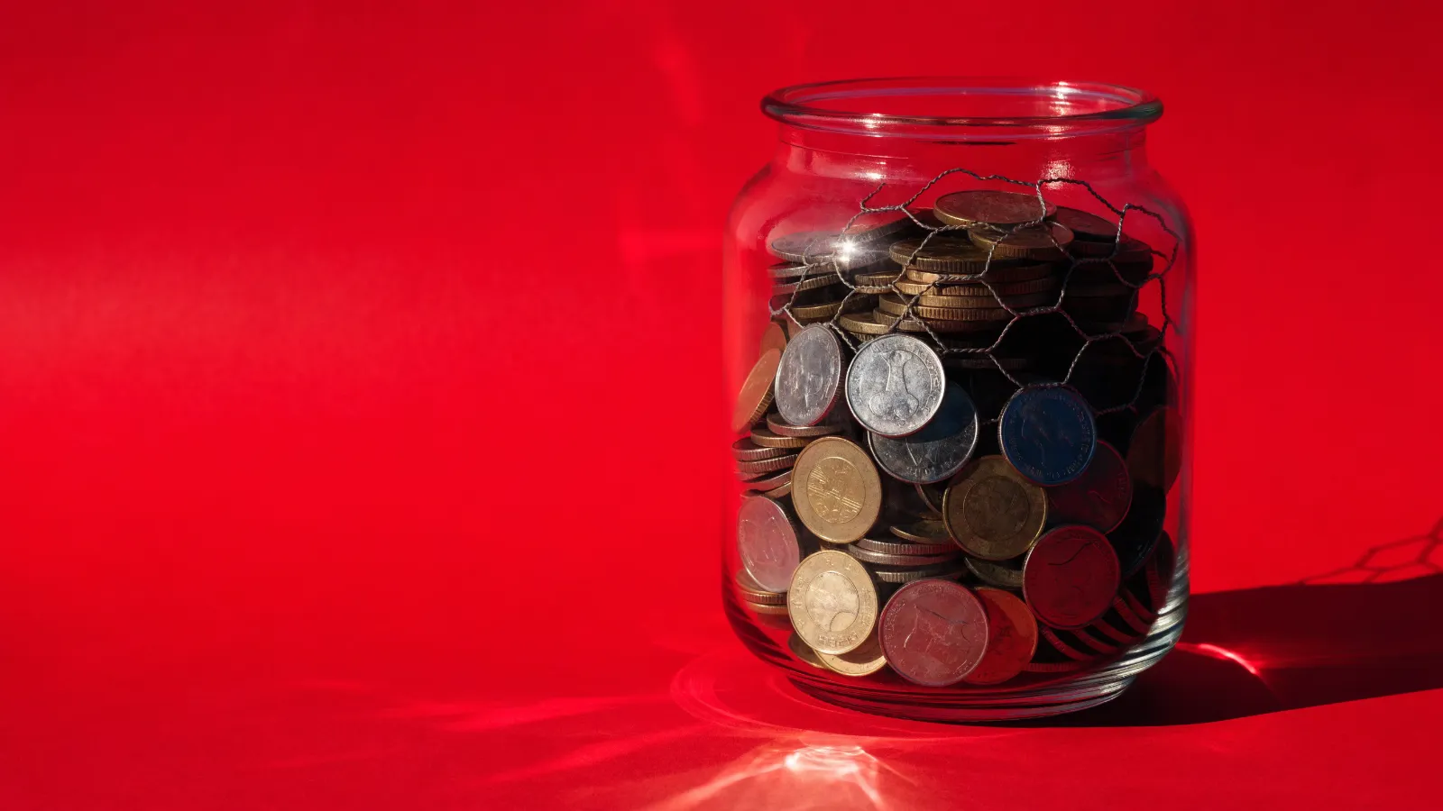 Un frasco de monedas con una parte de ellas encerrada en una jaula de malla, sobre fondo rojo